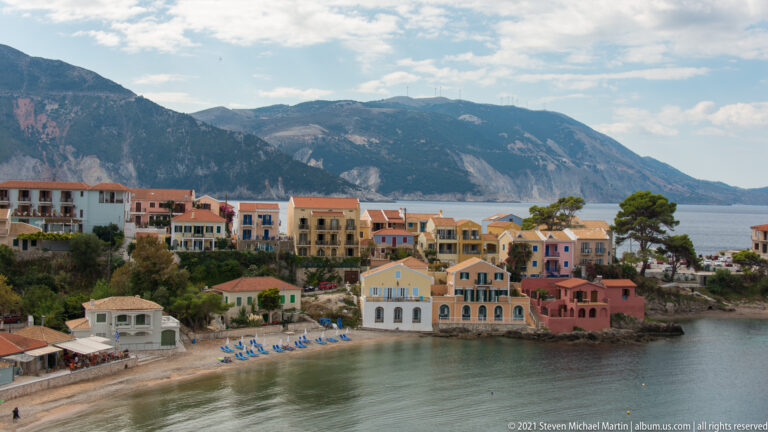 Pastel Assos buildings beside a narrow beach with blue lounge chairs, with a massive rocky mountain across the bay.