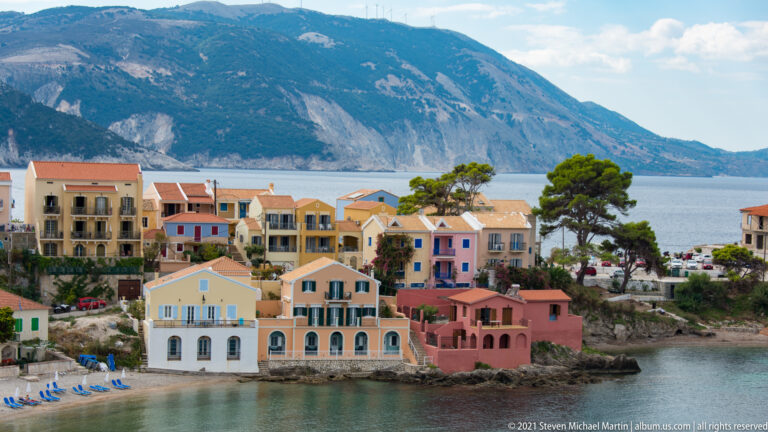 Elevated view of pastel waterfront houses and narrow beach in Assos, with a large mountain rising across the bay.