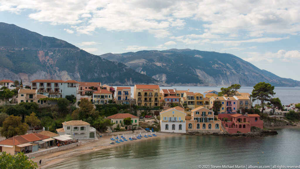 Wide view of Assos cove with pastel houses, narrow beach, blue lounge chairs, and mountain wall across the water.