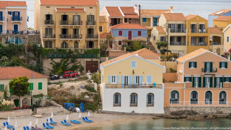 Yellow and peach Assos waterfront houses with shutters, balconies, arched lower windows, tiled roofs, and blue beach chairs nearby.