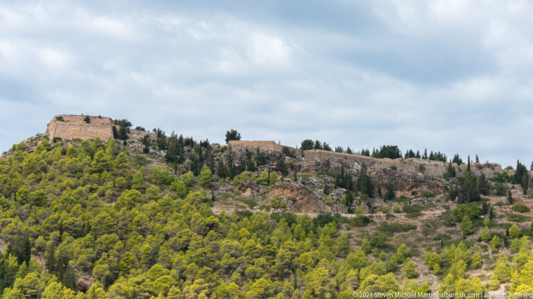 Distant stone fortification extending across a wooded ridge above Assos, with thick walls and exposed rocky slopes below.