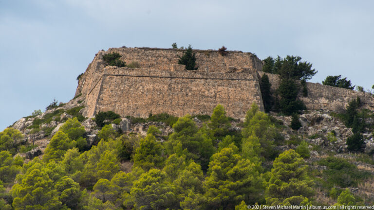 Large angular stone fortification with battered walls rising above a pine-covered rocky slope at Assos.