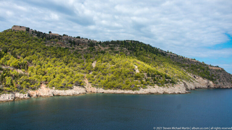 Wooded Assos peninsula descending to blue water, with pale stone fortification walls visible along the upper ridge.