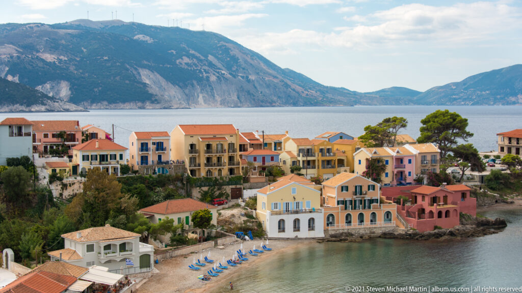 Elevated panorama of Assos with pastel waterfront buildings, narrow beach, blue lounge chairs, cove, and mountain beyond.