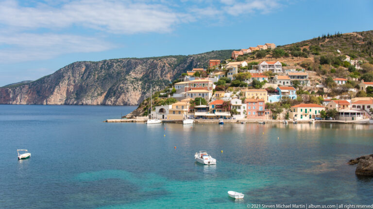 Pastel waterfront houses and small boats beside clear turquoise shallows in Assos harbor, with a rocky hillside behind.