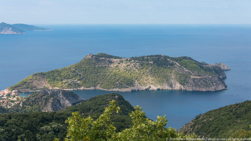Elevated view of the forested Assos peninsula, sheltered cove, blue sea, village edge, and pale fortification walls along the ridge.