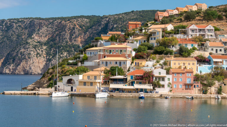 Pastel harbor-front buildings with balconies and terraces beside moored sailboats, backed by a steep rocky mountainside.