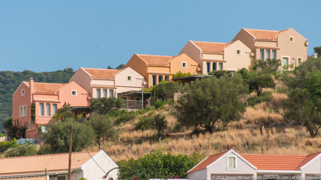 Row of pale stucco villas with red-tiled roofs and balconies on a dry hillside with olive trees.