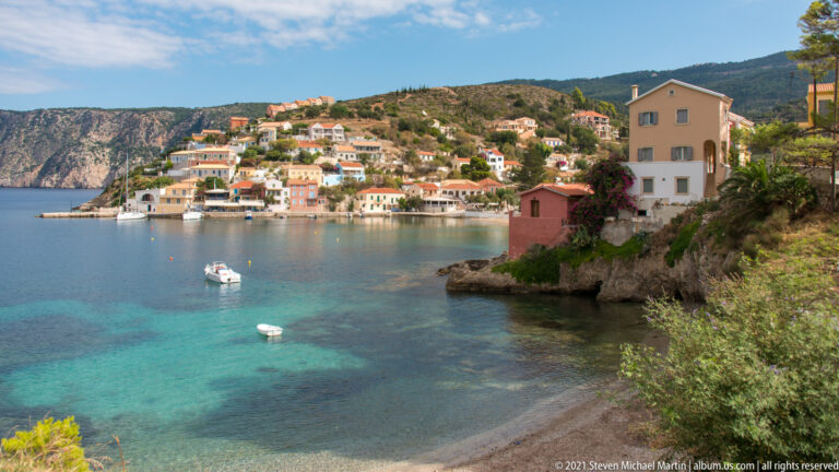 Assos village across turquoise bay water, with small boats, pastel houses, hillside homes, and a rocky mountain backdrop.