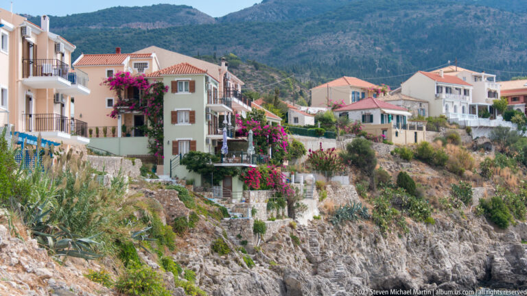 Assos houses with balconies and bougainvillea above a jagged rocky shoreline, with mountains behind.