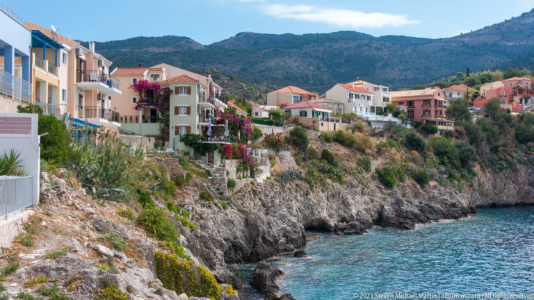 Oblique view of pale houses, balconies, gardens, and bougainvillea above rocky blue-water shoreline in Assos.