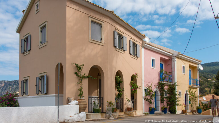 Peach house with rounded ground-floor arches, gray shutters, climbing plants, and a neighboring pink building on an Assos lane.