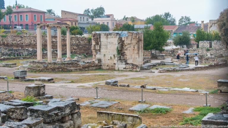 Roman Agora excavation with three standing columns, a large stone pier, low foundations, and neighborhood buildings beyond.
