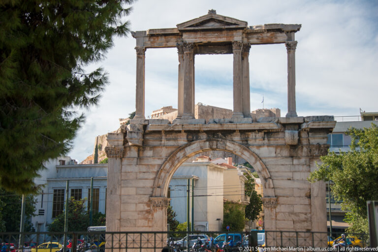 Hadrian's Arch with broad lower opening, upper columns, central pediment, weathered marble, and modern buildings behind.