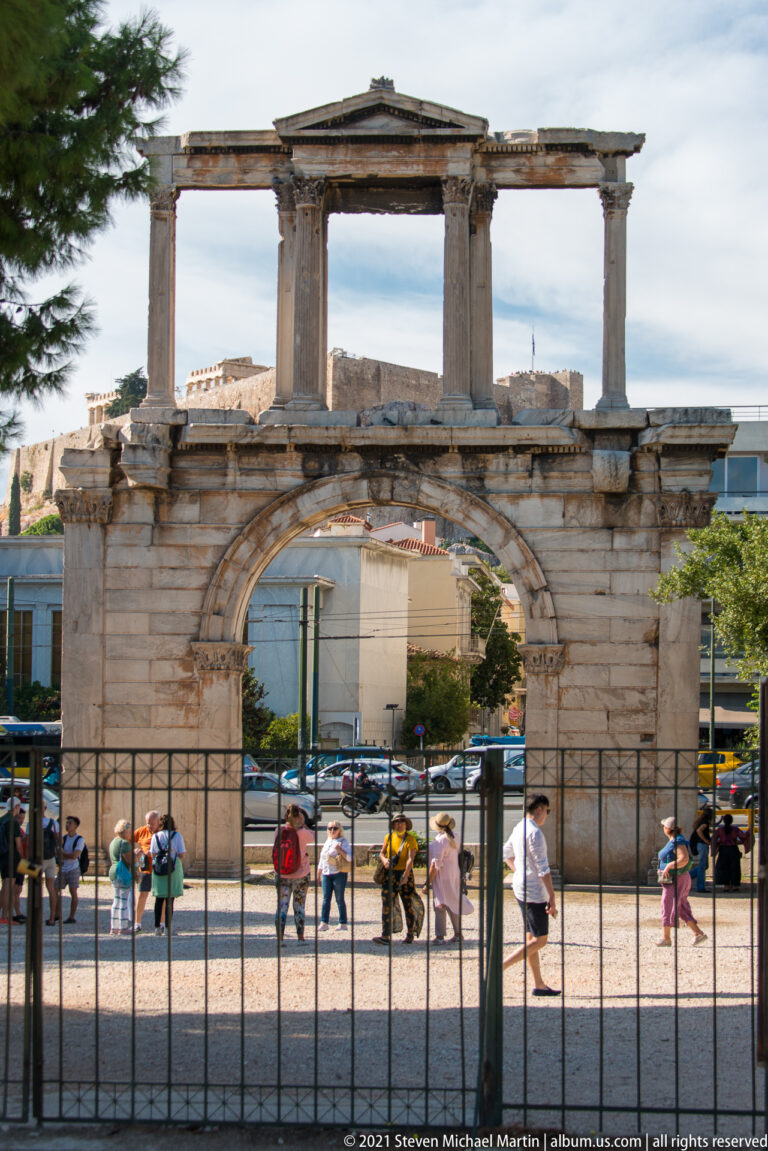 Hadrian's Arch behind a black fence with pedestrians walking through the paved urban space beyond.