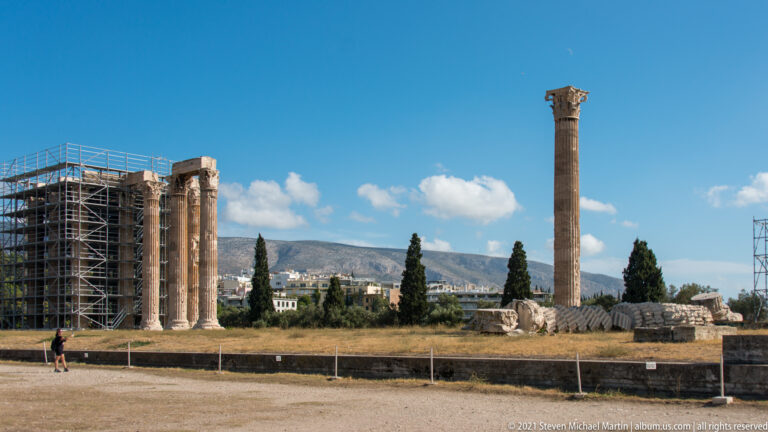 Wide Olympieion view with isolated Corinthian column, scaffolded column group, fallen drums, cypress trees, and mountains.