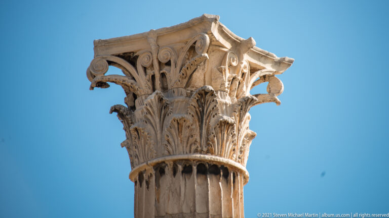 Close view of a weathered Corinthian capital with deeply carved acanthus leaves and volutes against blue sky.