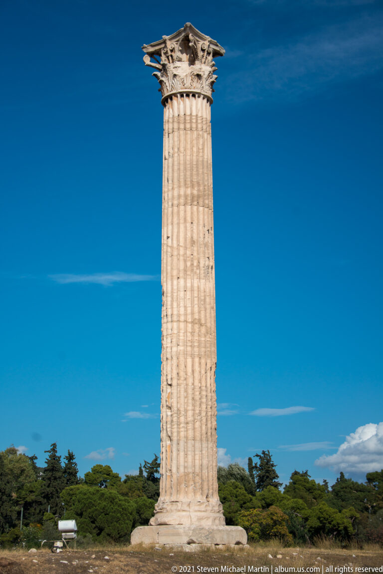 Full isolated Corinthian column of the Temple of Olympian Zeus rising above low trees against blue sky.