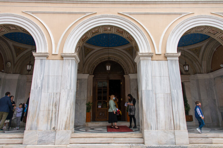 Three-arched marble entrance portico of Athens Metropolitan Cathedral with blue painted vaults, doors, steps, and visitors.