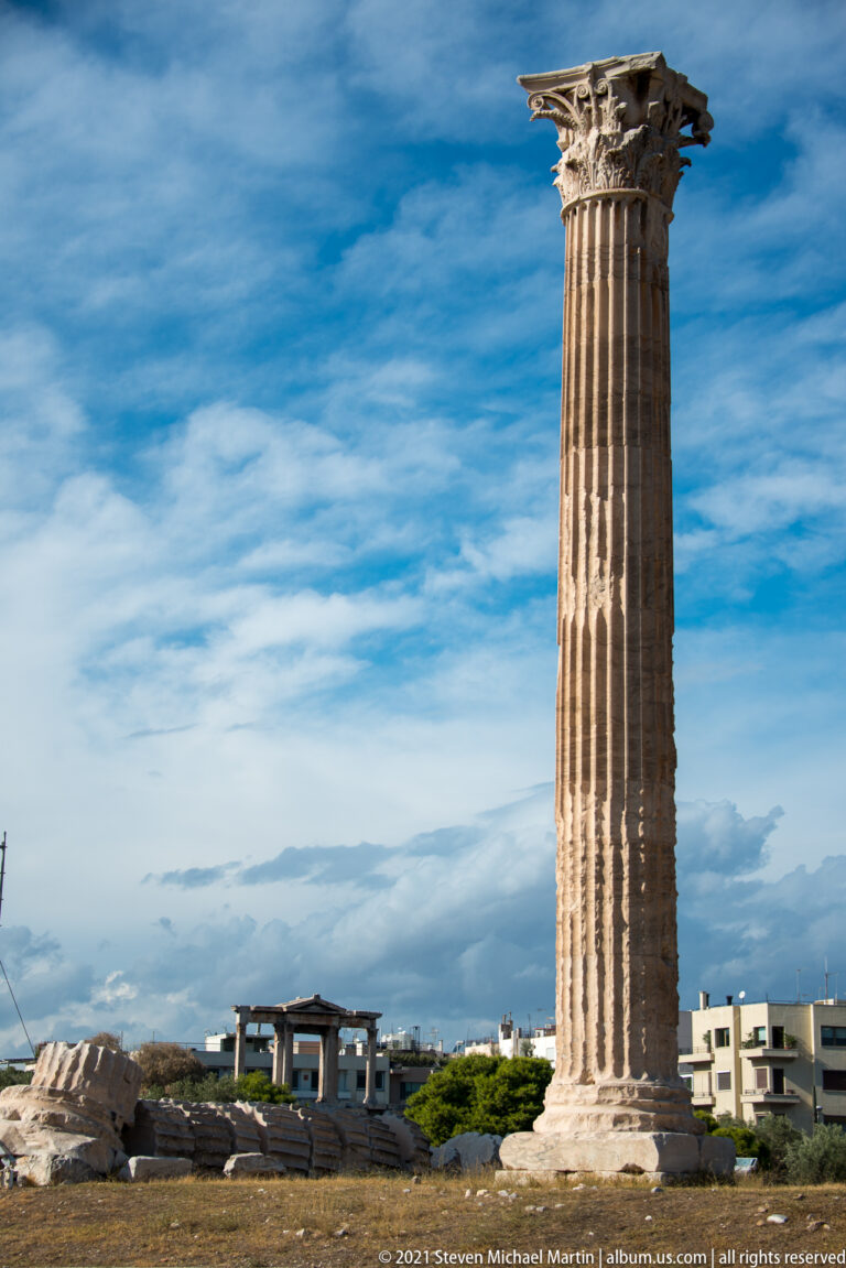 Single weathered Corinthian column of the Temple of Olympian Zeus standing above fallen blocks and modern Athens.