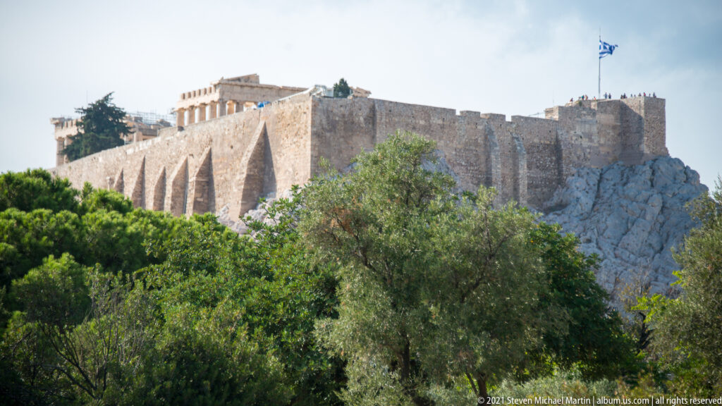 Acropolis walls and Parthenon rising above green trees, with a Greek flag flying at the summit.