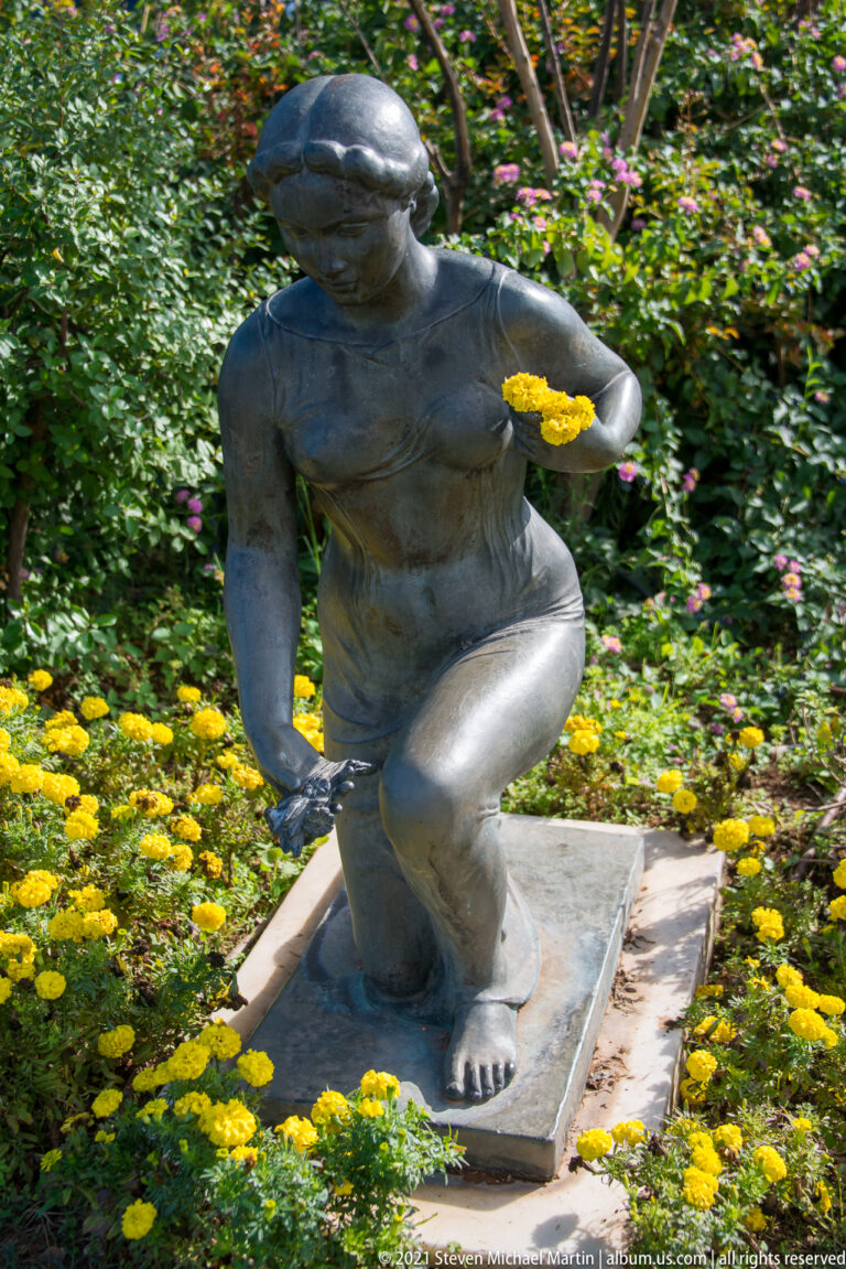 Crouching dark bronze nude figure reaching toward a yellow flower amid dense green foliage and low yellow blossoms.