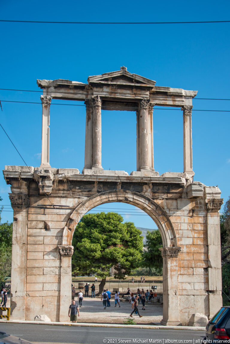Frontal Hadrian's Arch with upper colonnade and central pediment, pedestrians visible through the large lower opening.