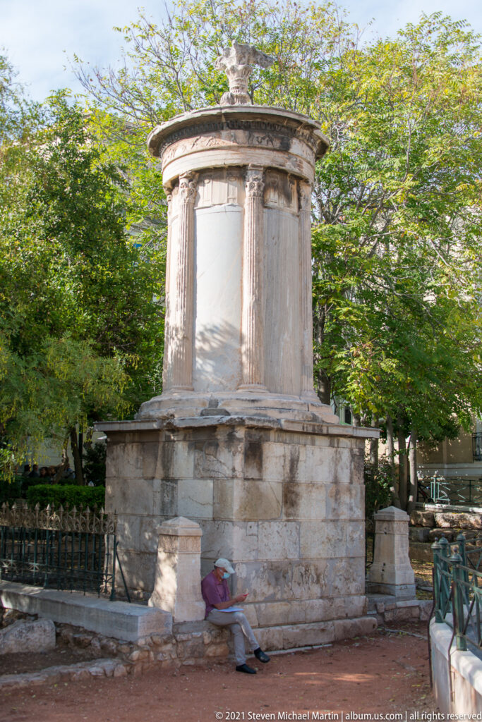 Wider view of the Choragic Monument of Lysicrates among trees in Plaka, showing its square base and circular Corinthian upper structure.