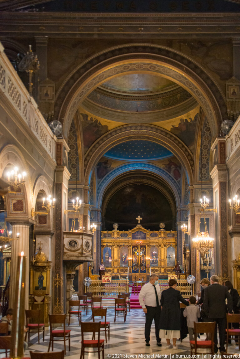 Ornate nave of Athens Metropolitan Cathedral facing a gilded iconostasis beneath painted arches, chandeliers, and a coffered ceiling.