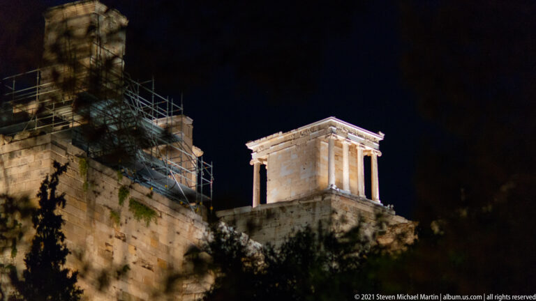 Oblique night view of the illuminated Gate of Athena Archegetis in the Roman Agora, with Doric columns and adjacent conservation scaffolding.