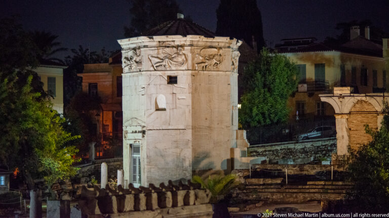 Floodlit Tower of the Winds with sculpted frieze and dark roof above Roman Agora ruins at night.