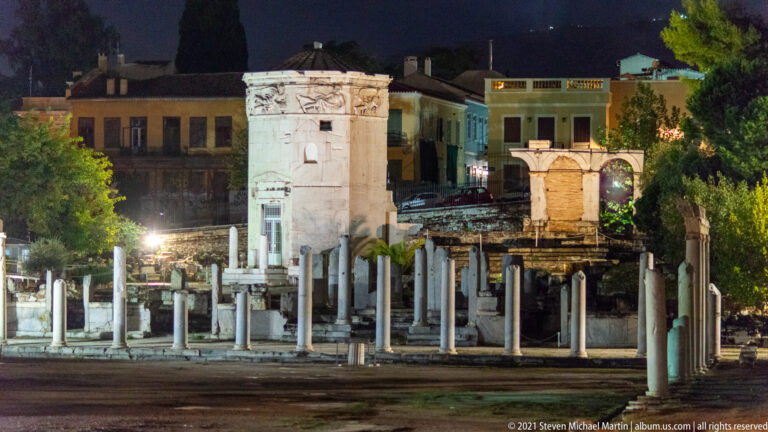 Wide night view of Roman Agora columns and ruins with the illuminated Tower of the Winds rising behind.