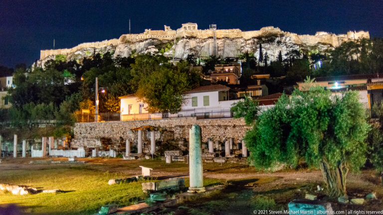 Roman Agora ruins in the foreground beneath the brightly illuminated Acropolis ridge at night.