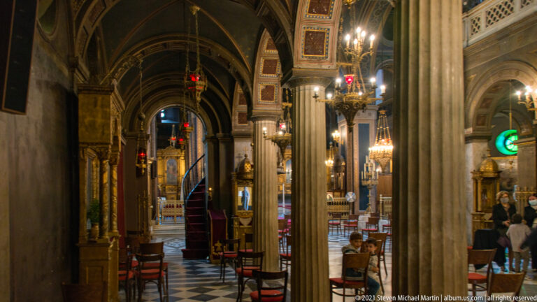 Columned side aisle inside Athens Metropolitan Cathedral with icons, chandeliers, painted arches, chairs, and a black-and-white floor.