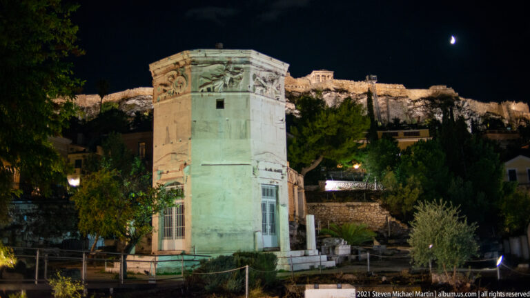 Floodlit Tower of the Winds below the illuminated Acropolis ridge with a crescent moon in the night sky.