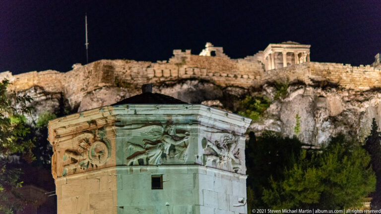 Close view of winged relief figures on the Tower of the Winds with the illuminated Acropolis ridge behind.