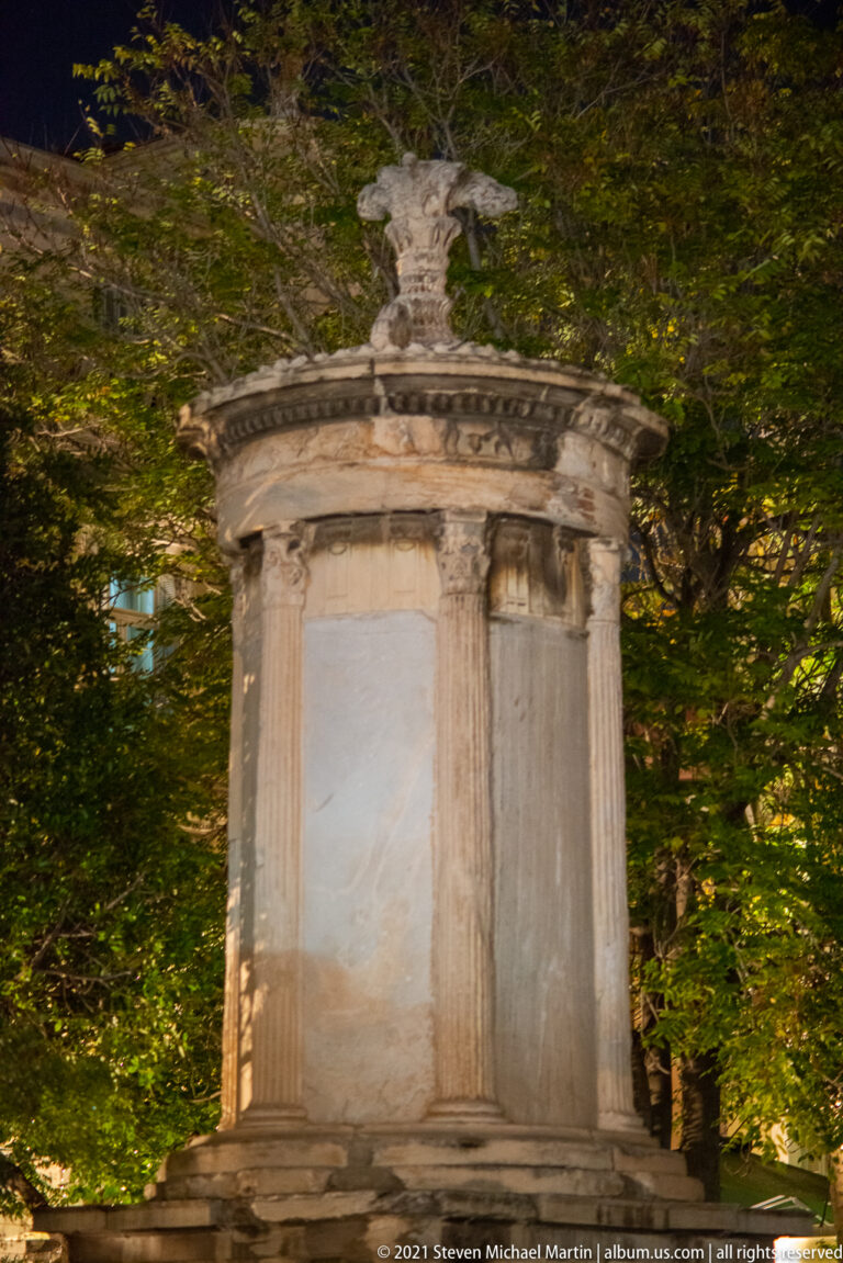 Close view of the Choragic Monument of Lysicrates among trees, showing engaged Corinthian columns, marble wall panels, frieze, and finial.