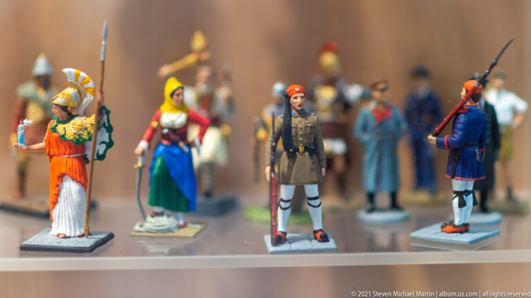 Painted miniature Greek figures in ancient, folk, and military costumes arranged in rows on a display shelf.