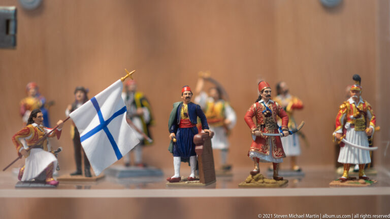 Miniature Greek fighters in traditional dress with rifles and swords, including a flag bearer carrying a blue-cross flag.