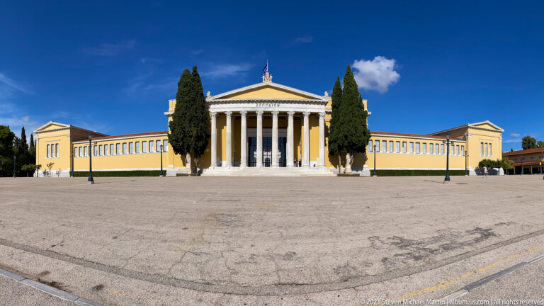National Archaeological Museum of Athens with central white Ionic portico, yellow wings, cypress trees, and broad forecourt.