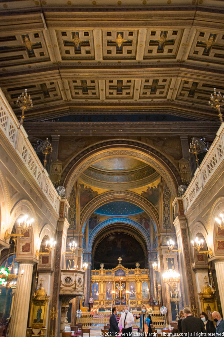 Wide interior view of Athens Metropolitan Cathedral with coffered ceiling, painted arches, chandeliers, marble columns, and gilded iconostasis.