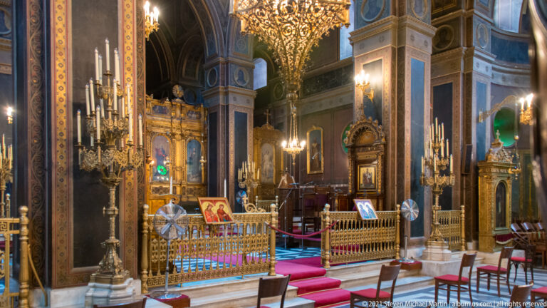 Gilded icon stands, candleholders, carved sanctuary rail, framed icons, and marble piers inside Athens Metropolitan Cathedral.