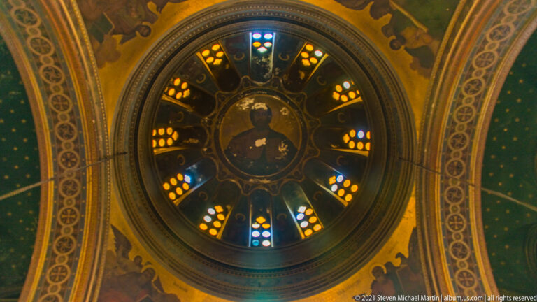 Circular ceiling medallion of Christ ringed by colored glazing within a richly painted blue-and-gold cathedral vault.