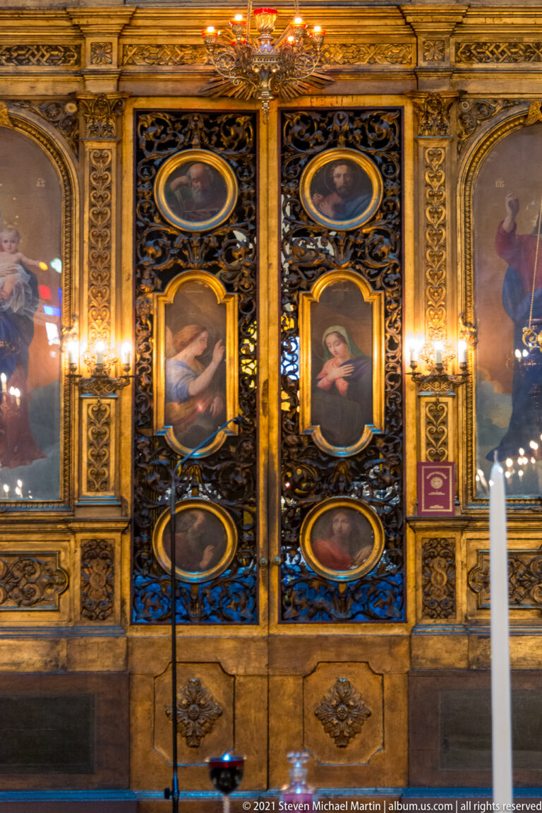 Dark carved screen with gilded trim and stacked oval and rectangular painted devotional panels inside Athens Metropolitan Cathedral.