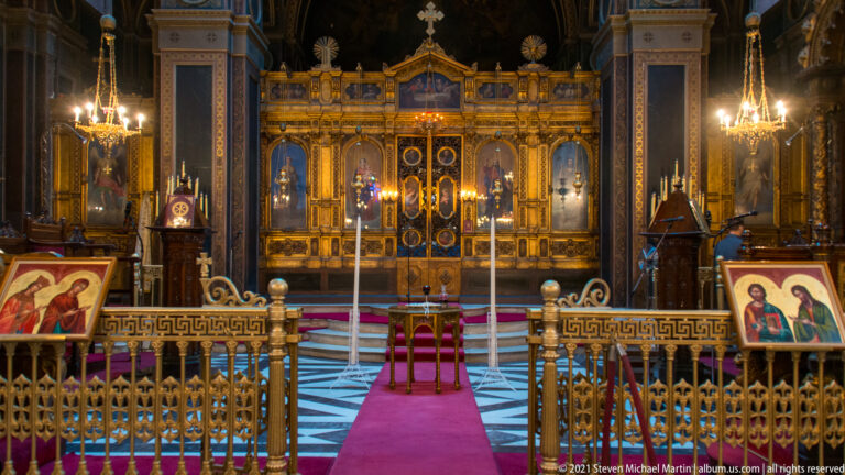 Frontal gilded iconostasis with royal doors, icons, lamps, brass rail, and red carpet in Athens Metropolitan Cathedral.