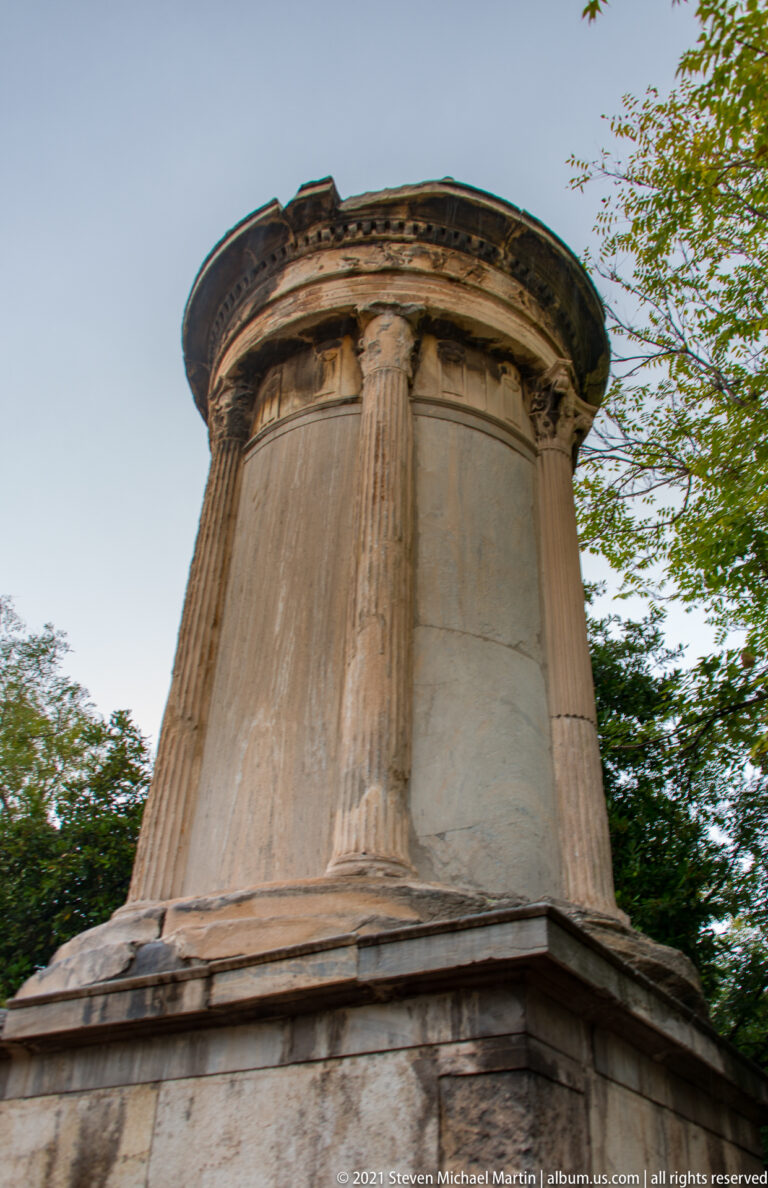 Low-angle view of the circular Choragic Monument of Lysicrates with fluted Corinthian columns, sculpted frieze, and trees.