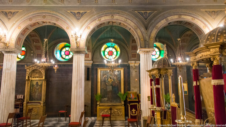 Three arched bays with circular stained glass, marble columns, icons, and lamps inside Athens Metropolitan Cathedral.