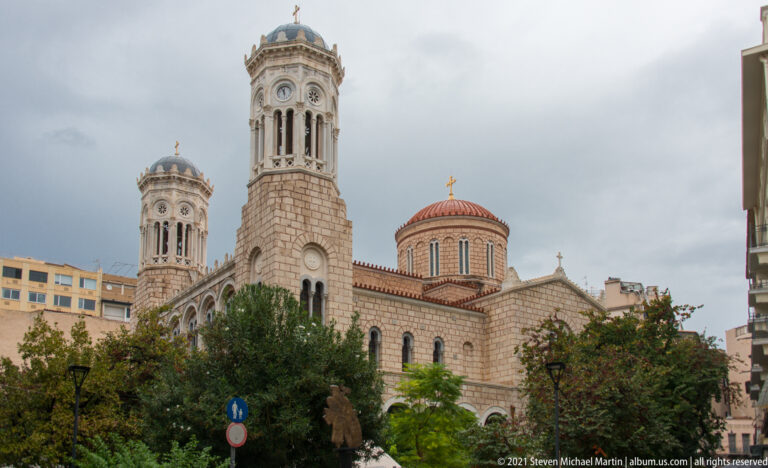 Athens Metropolitan Cathedral exterior with twin bell towers, patterned masonry, arched windows, red-tiled dome, and trees.