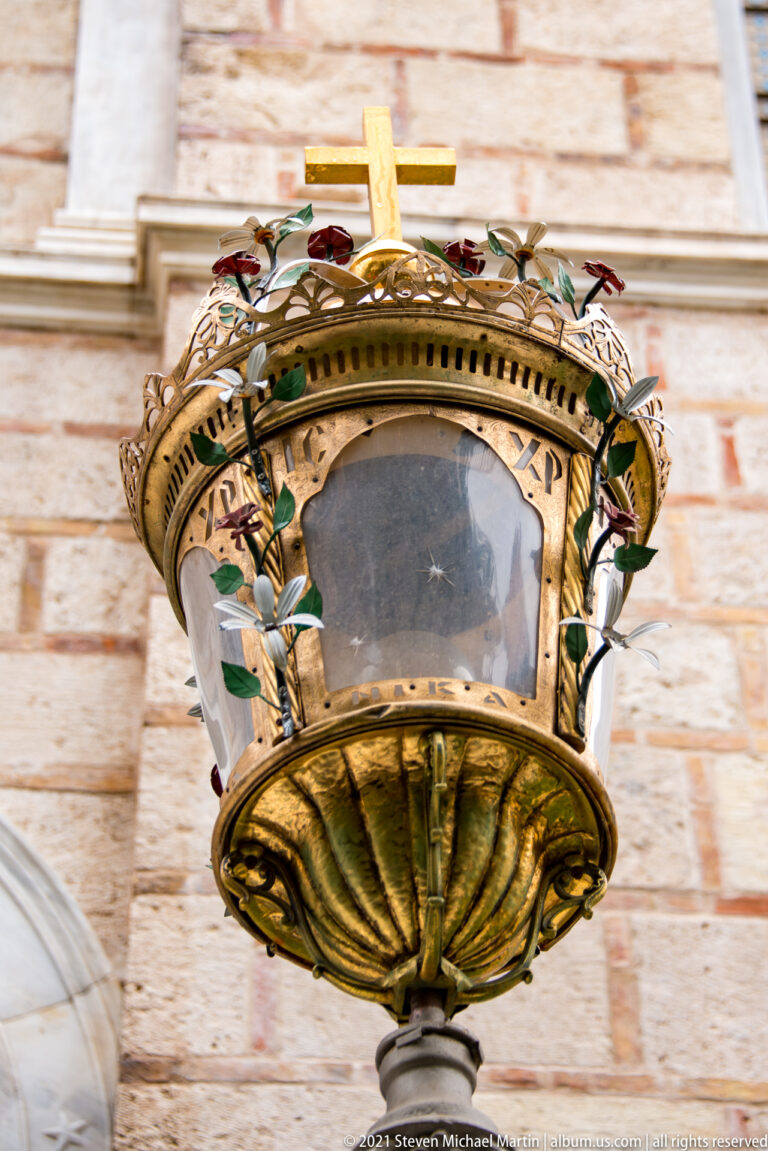 Ornate suspended metal church lamp with cross, leafy decoration, and colored accents against pale stone masonry.