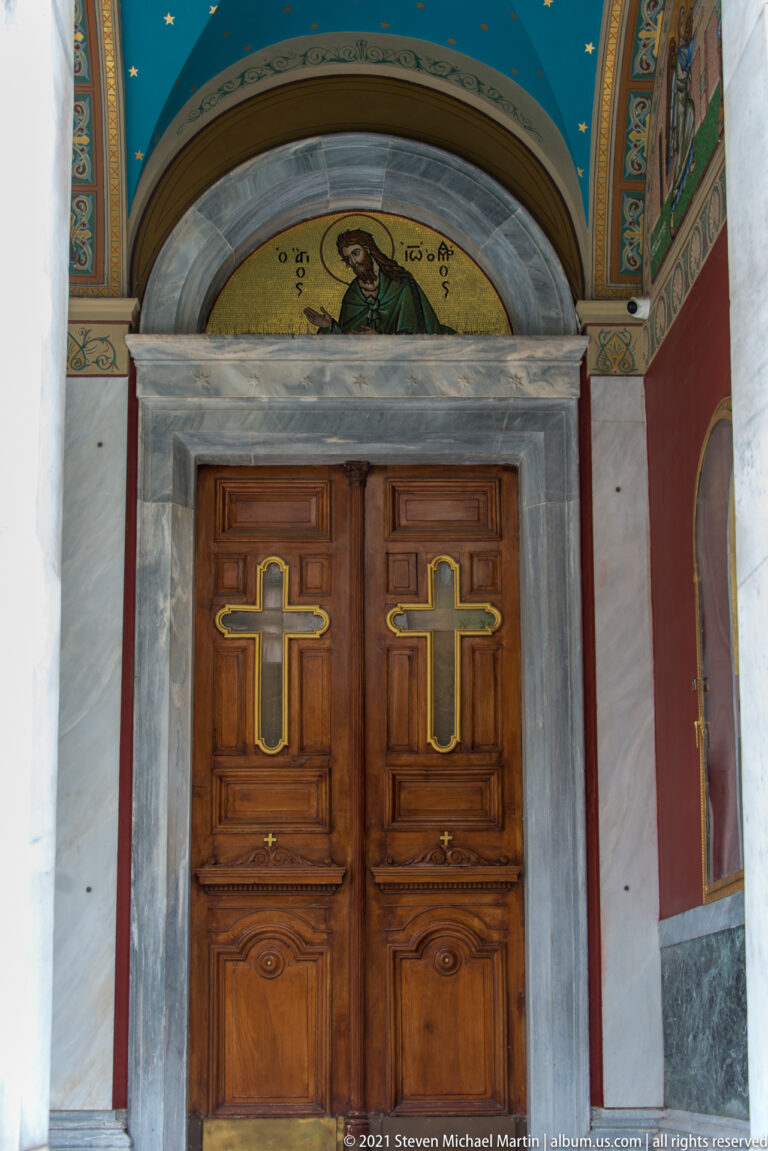 Wooden cross-paneled doors beneath a mosaic of Christ and blue painted vault at Panagia Chrysospiliotissa in Athens.